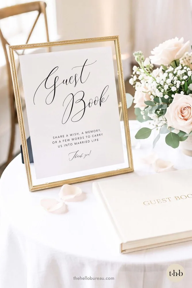 Elegant script guestbook sign in a gold frame beside blush roses, baby’s breath and a cream guestbook