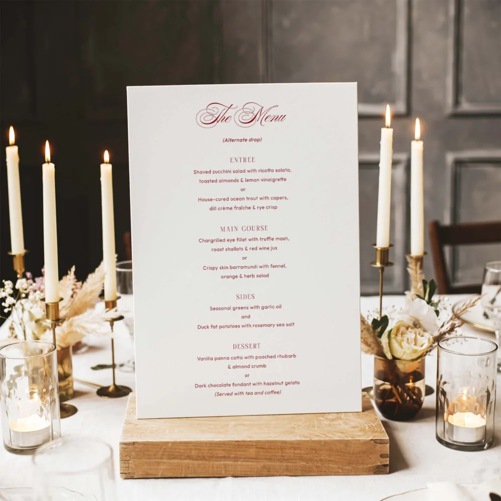 Individual Arden wedding menu template featuring romantic red script and a clean serif layout. Shown in a candlelit reception setting on a rustic timber block. Ideal for an elegant alternate drop dinner service.