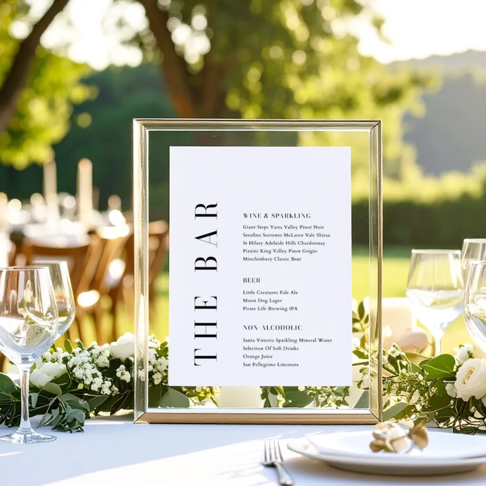 Modern Brooklyn bar menu sign in a landscape gold frame, featuring bold vertical typography. Displayed on an outdoor reception table with white florals and greenery for a contemporary garden wedding.
