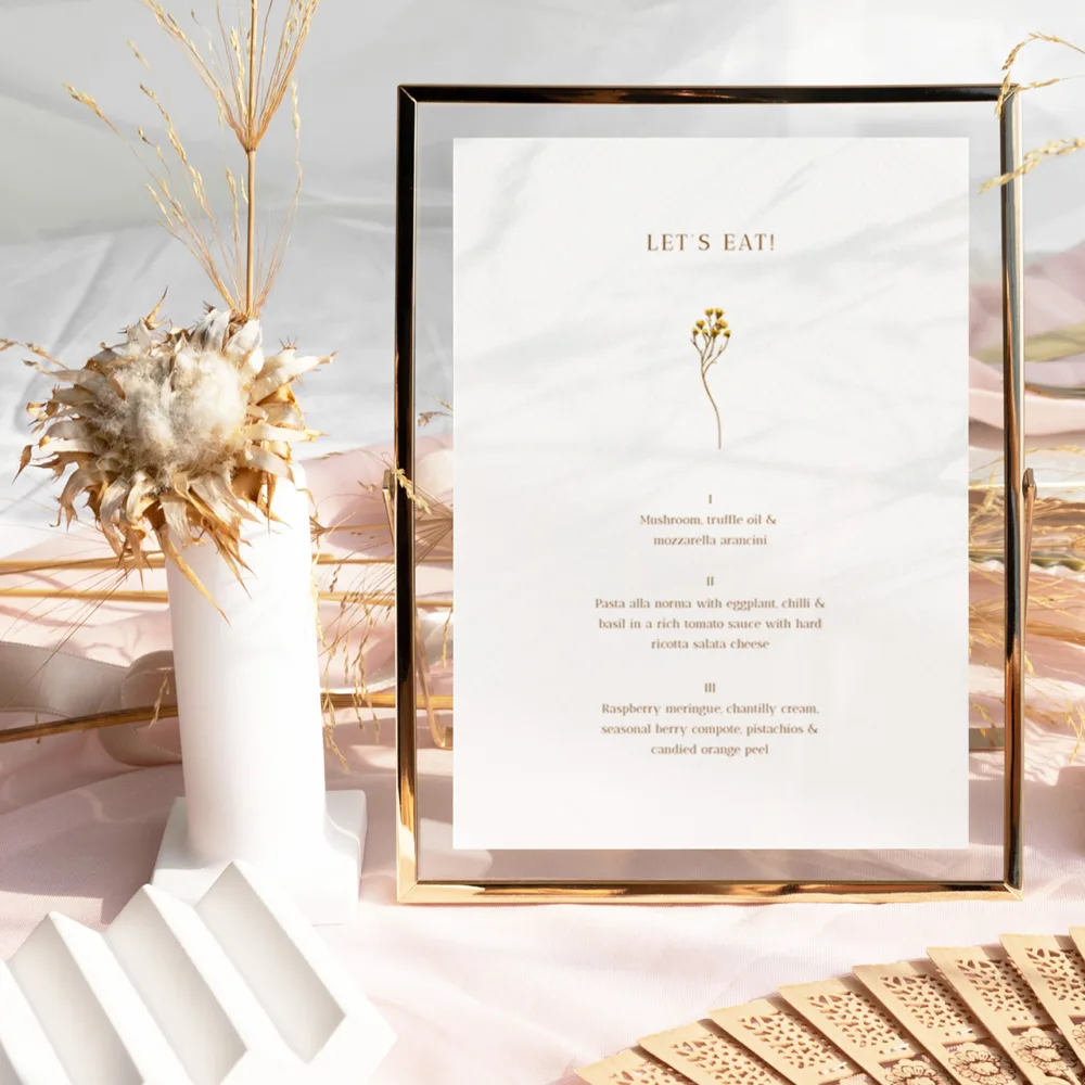 Minimalist wedding menu card with a delicate botanical illustration, displayed in a modern gold floating frame. The Goldfields design by The Hello Bureau is perfect for a sophisticated, nature-inspired table setting.