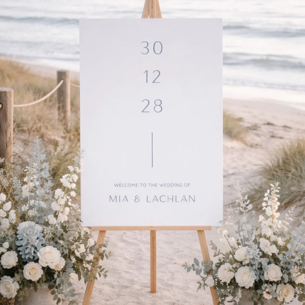 Coastal-inspired wedding welcome sign featuring soft blue typography and a minimalist vertical layout. Shown on a beach with sand dunes and white florals, the Laguna design is a stress-free DIY choice for a seaside ceremony.