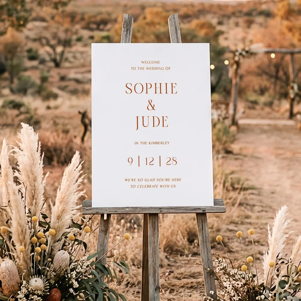 Earthy, terracotta-toned wedding welcome poster displayed on a rustic timber easel in an outback desert setting. The Terra design by The Hello Bureau is inspired by the Kimberley and is ideal for warm, boho-themed celebrations.