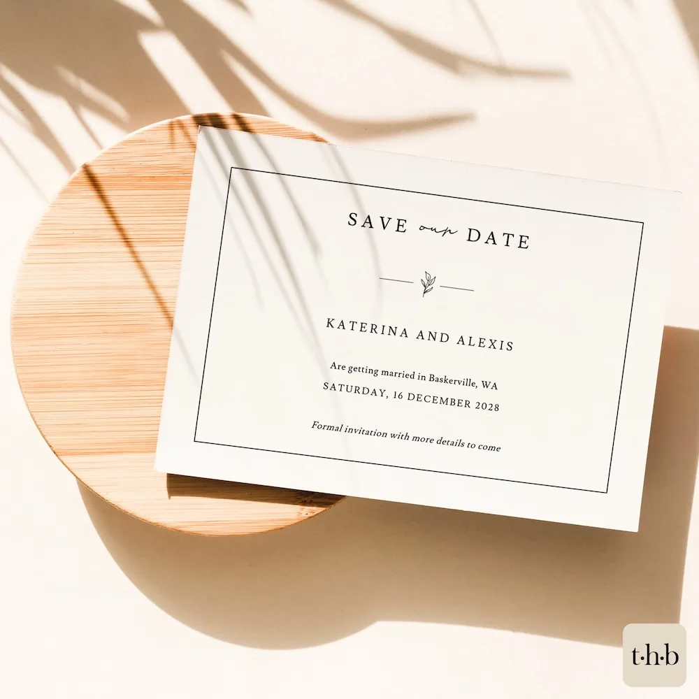 A white save the date card with an elegant design on a wooden plate.