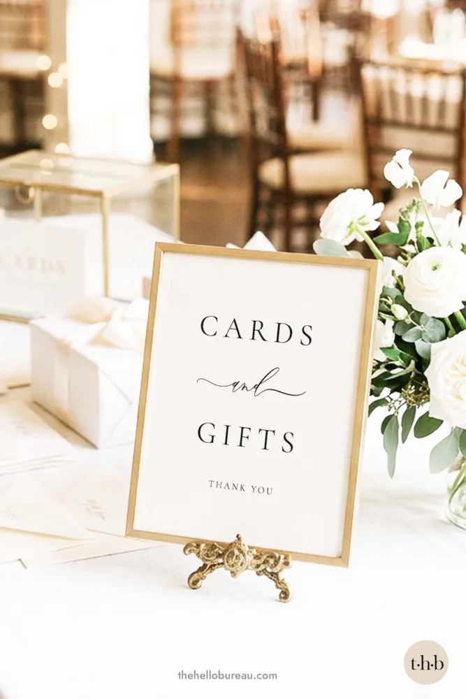 A classic and formal wedding cards and gifts sign by The Hello Bureau, displayed in a thin gold frame on a white table with white flowers and gift boxes.