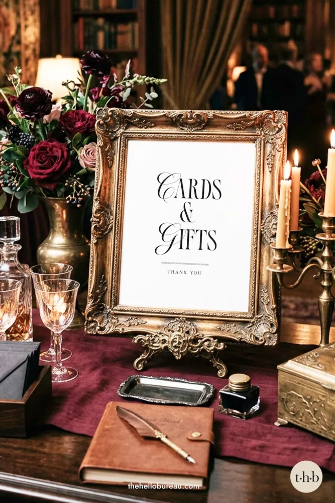 An Old Money and Editorial style cards and gifts sign from The Hello Bureau, styled in an ornate vintage gold frame on a burgundy tablecloth with dark roses and candles.