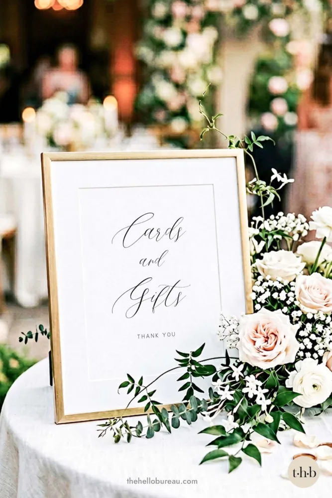 An elegant script wedding cards and gifts sign from The Hello Bureau, displayed in a gold frame on a reception table surrounded by soft pink roses and white baby's breath.