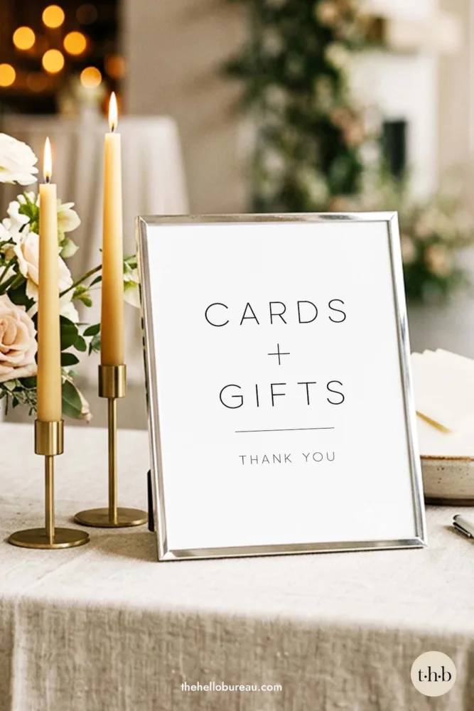 A minimal sans serif cards and gifts sign by The Hello Bureau, styled in a sleek silver frame on a linen tablecloth next to gold candle holders and tall tapers.
