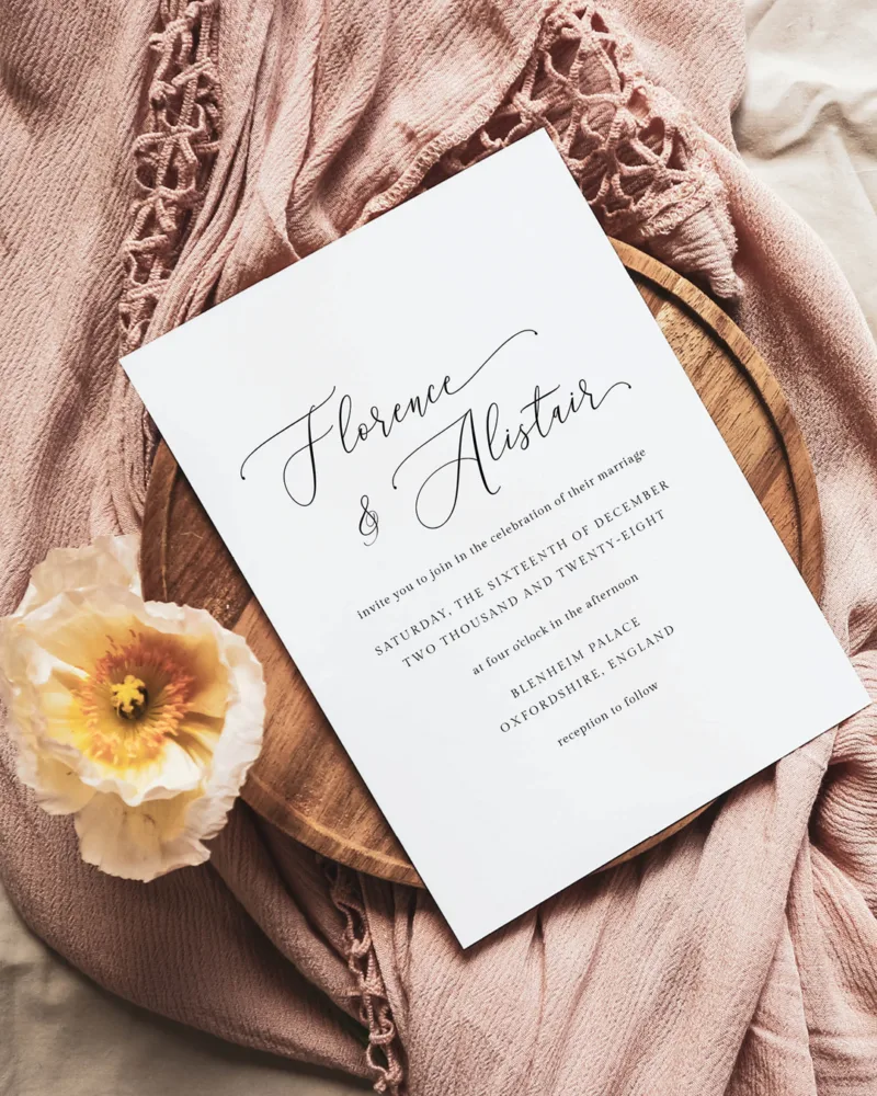 A wedding invitation card on a pink linen background with a flower.