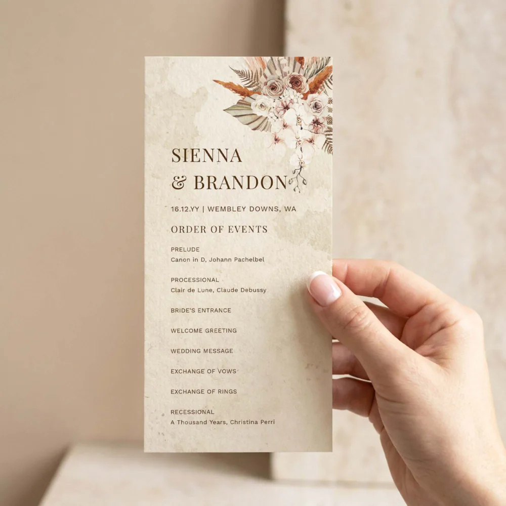 Hand holding a DIY boho wedding ceremony program template with dried floral arrangement, earthy watercolour texture and order of events listing.