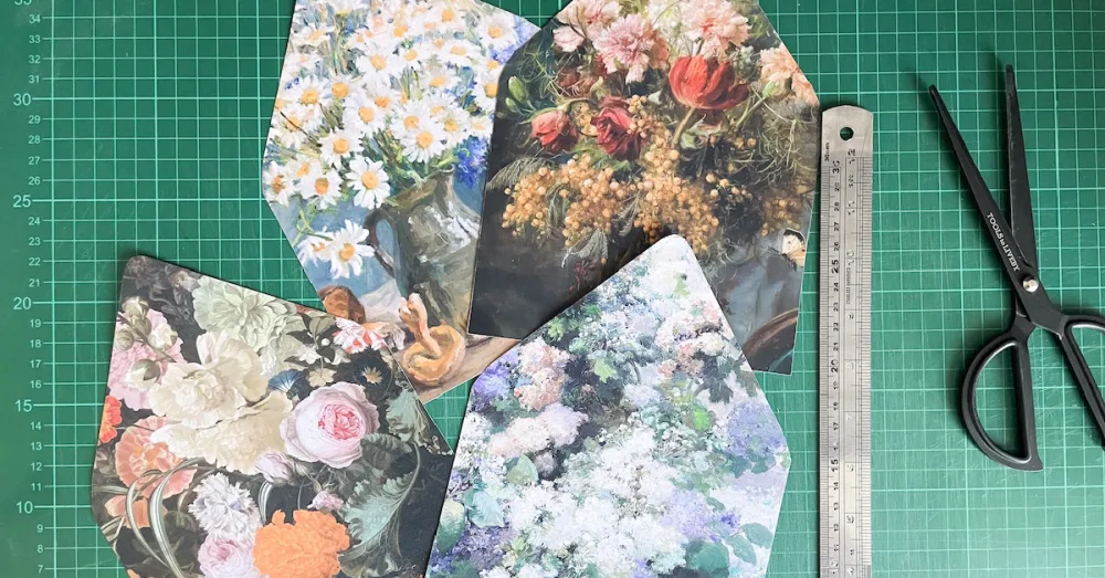 Four fine art floral envelope liners cut out and laid on a green cutting mat with a ruler and scissors.