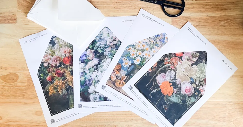 Four free printable A7 envelope liner templates in fine art floral designs, printed on A4 paper with blank envelopes and scissors on a wooden table.